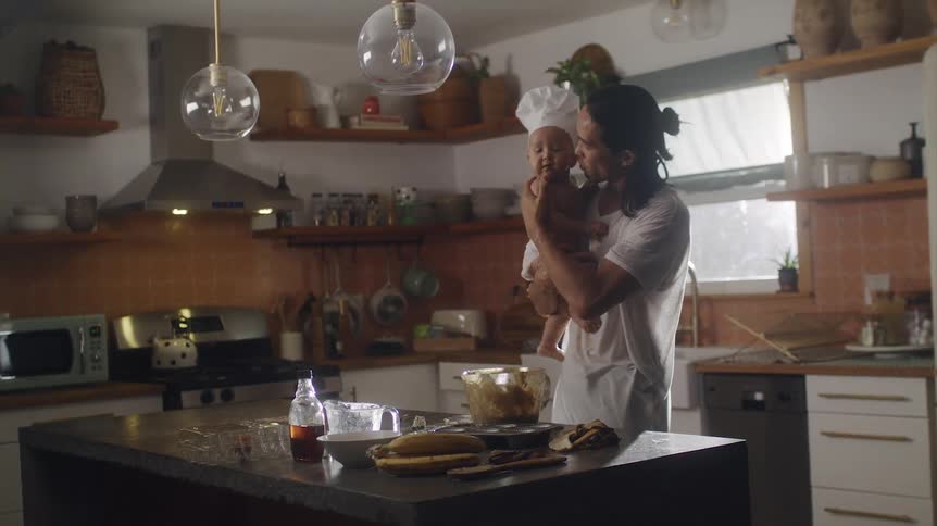 Father picks up child wearing a chef hat as he stand cooking at the kitchen island