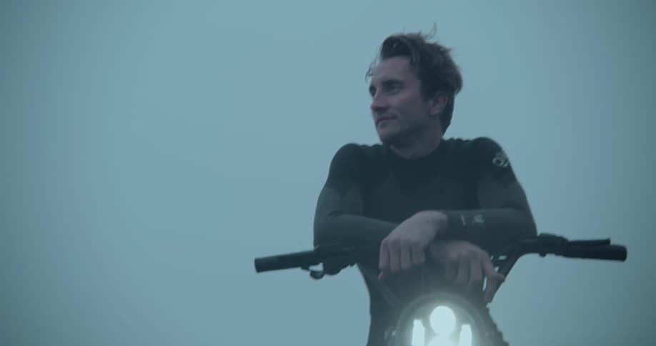 Man wearing a wetsuit sitting at his motorcycle resting his arms in the handlebar