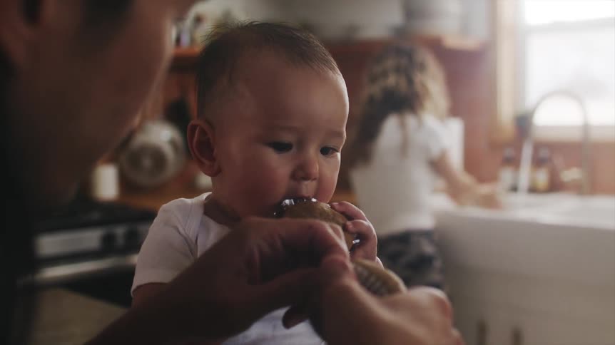 A man eats muffins with his baby while his partner washes dishes
