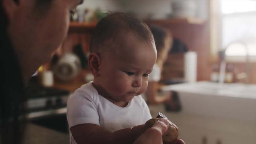 A baby tries to eat a muffin liner as a man tries to feed him pieces of a muffin