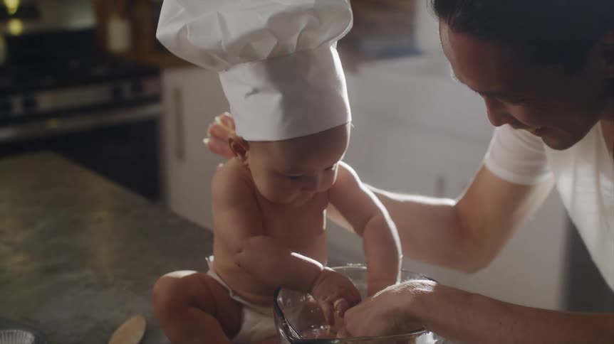 Father with long hair white shirt and apron plays dough in bowl with baby son wearing chef hat and apron sitting on kitchen island in the kitchen