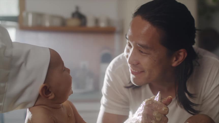 An asian man is looking at his baby wearing a chefs hat and kisses him and smiles as he holds his hand