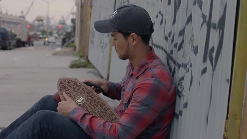 A young man sitting on the sidewalk is adjusting the wheel on a skateboard with a tool