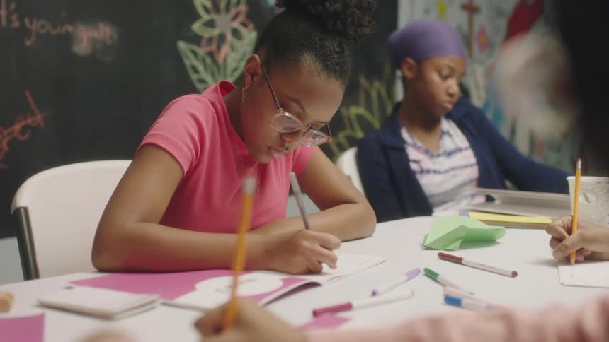 Girls color and draw at a long table in a classroom