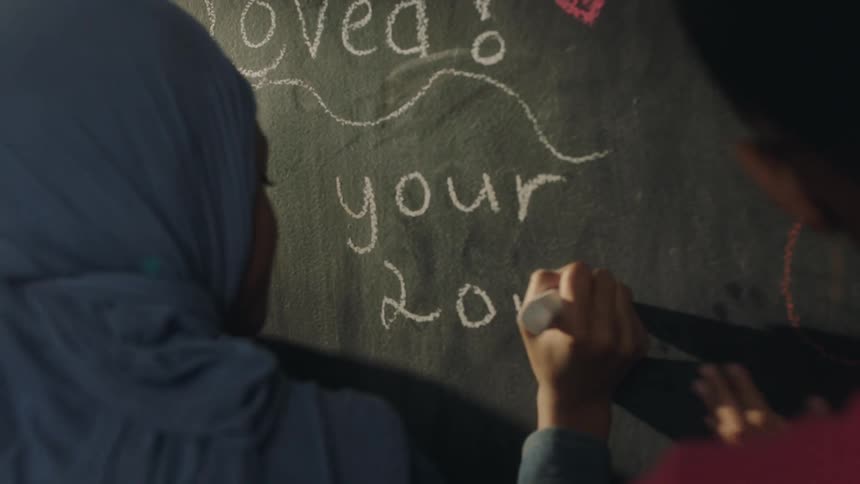 A pair of young women write on a blackboard using large pieces of chalk