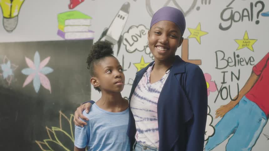 A young woman embraces a girl and smiles into the camera while the girl glares at her