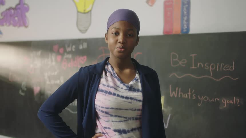 A young woman stands in a classroom with her hand on her hip while looking into the camera