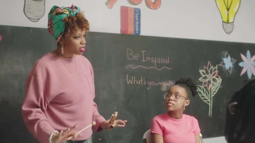 Woman in a pink sweater talks while standing in front of a chalkboard as a teenager listens while sitting in a chair
