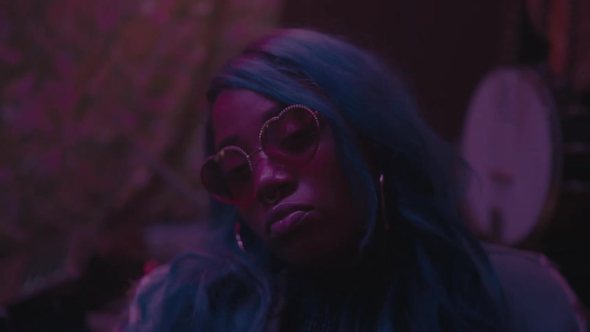 Black woman with blue hair and wearing heart shaped sunglasses looks contemplative