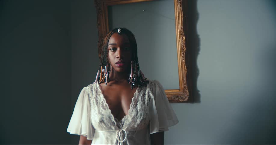 Woman with dreadlocks and white dress stands in front of wall with picture frame