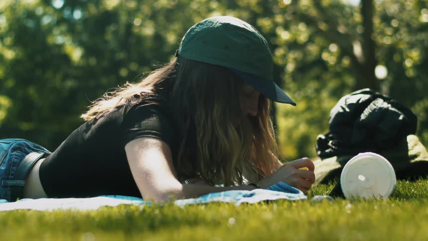 A woman participates in a video chat using a smartphone resting against a cup while lying on a blanket in a field during the day