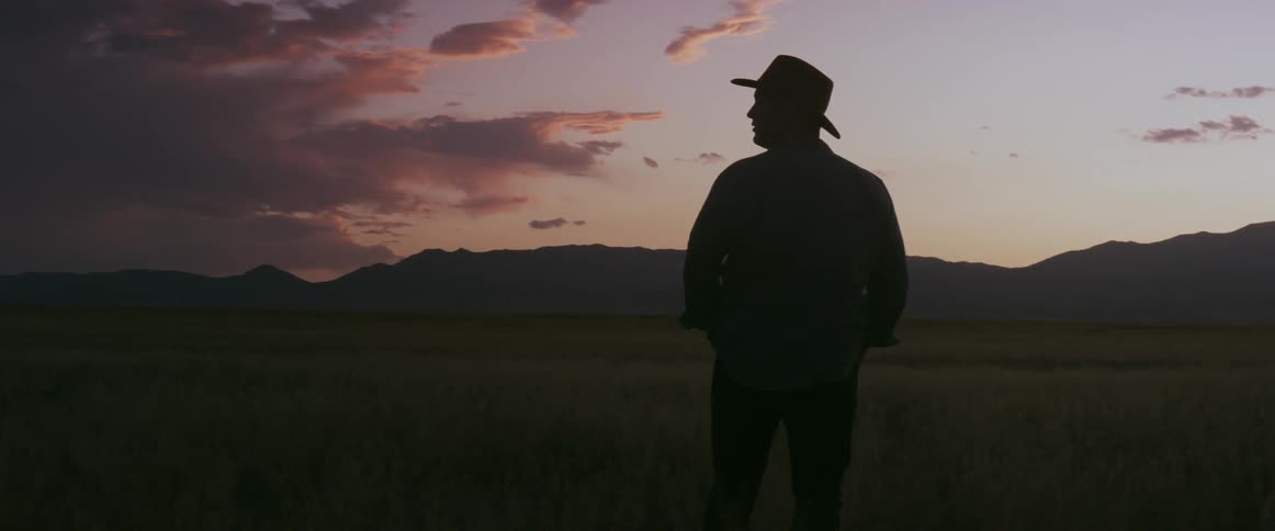 Man wearing cowboy hat stands in field looking toward mountains on horizon under red tinted clouds