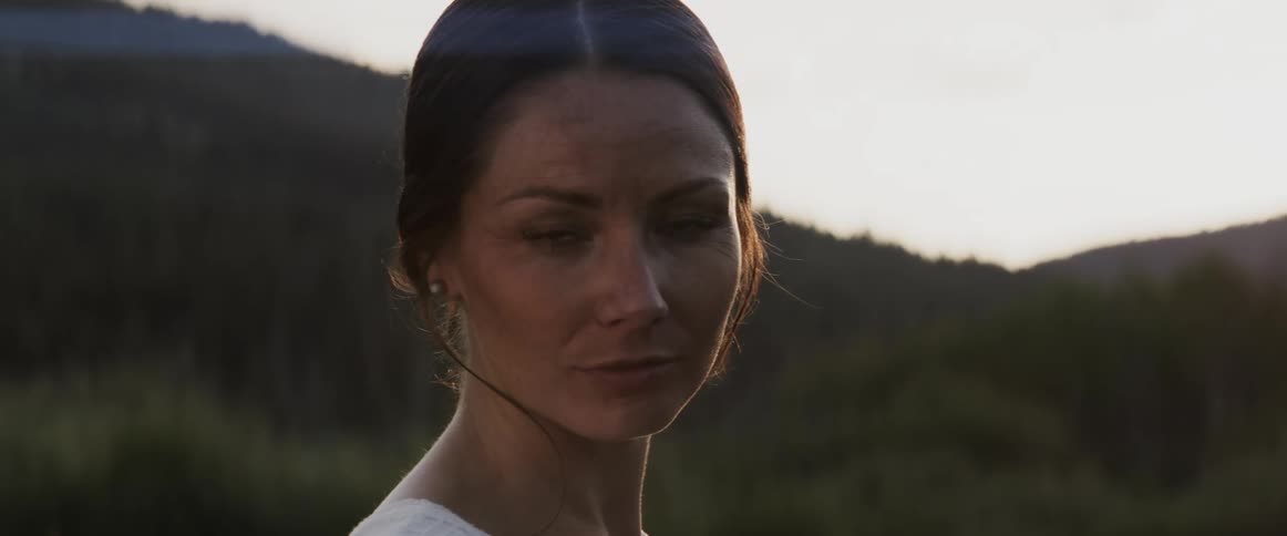 A woman gazes into the camera while standing in a hilly countryside during the day