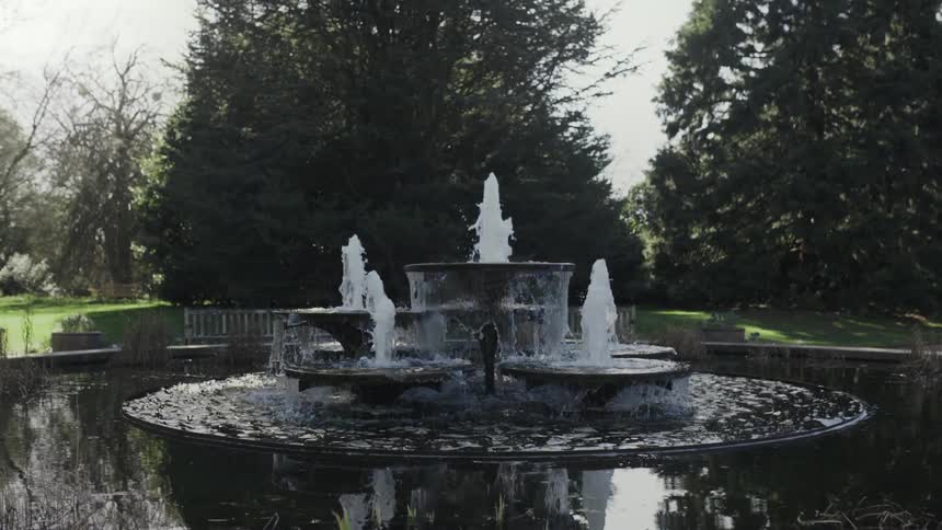 A water fountain flowing in a park with pool surface reflecting trees and sky
