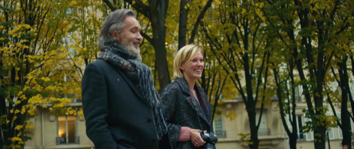 A man is walking through a park with a blond woman holding a camera
