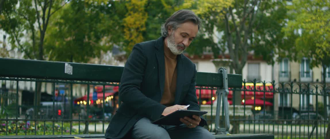 A man is sitting on a park bench using an electronic tablet