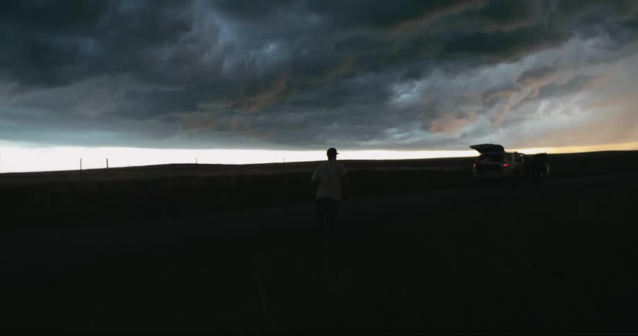 A man runs through the grass toward a parked car with door and hatch open while storm clouds gather above