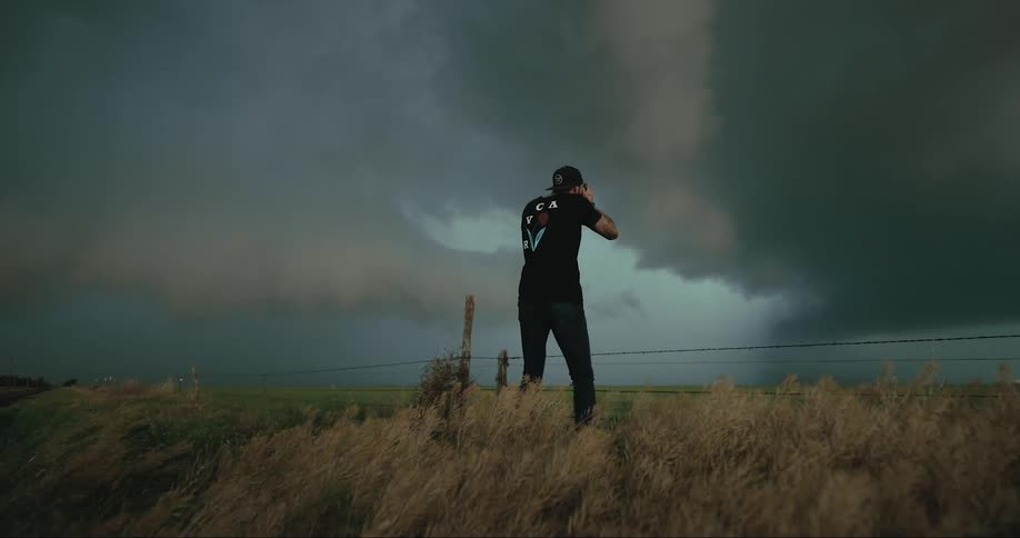 Man wearing a black hat black shirt and black pants takes a photo of a dark funnel cloud on a windy day