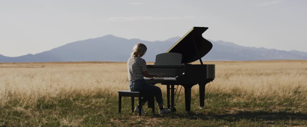 A woman sitting on a bench playing a piano in a field with hills on the horizon