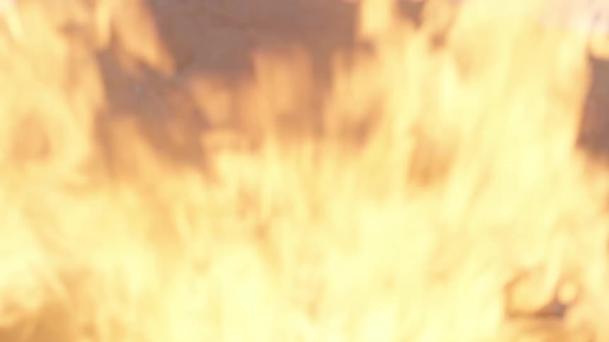 A man sits on a rocky hillside while lost in thought as a bonfire is ignited nearby