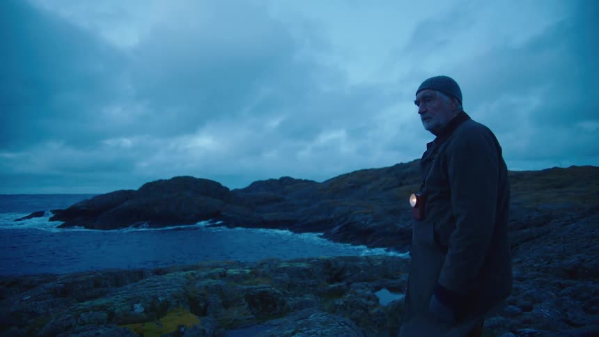 Man with wooly hat and overcoat stands and watches sea on the coast at dusk