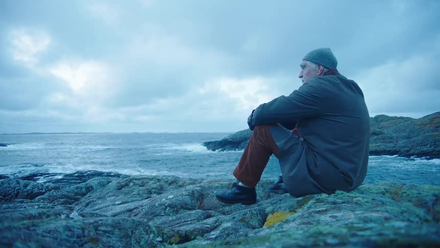 Elderly man with gray wooly hat overcoat brown pants and black shoes sits on rock and watches sea on cloudy day on the coast