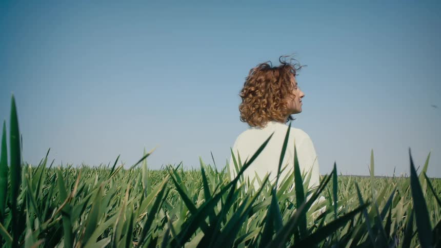 A woman sits in a grassy field and looks up at the sky as an insect flies past her