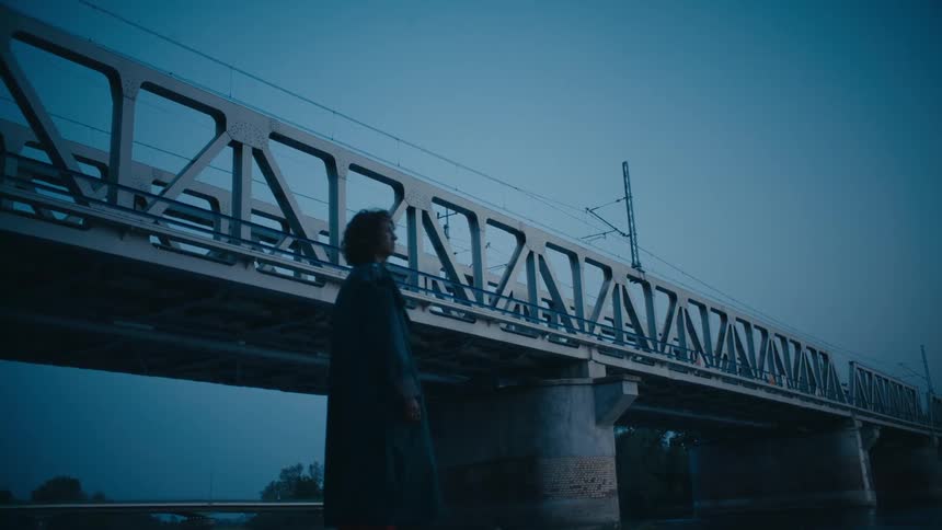 A person standing on a riverbank near a railroad bridge