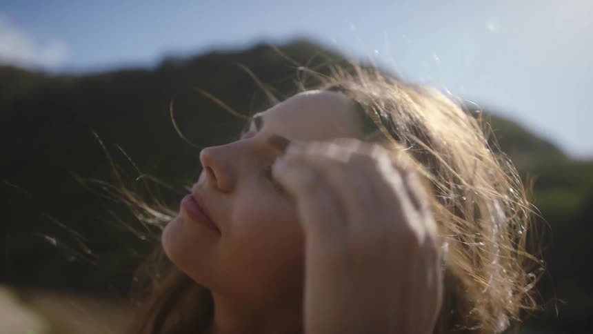 A woman runs her hand through her hair while standing outdoors with her eyes closed on a sunny day