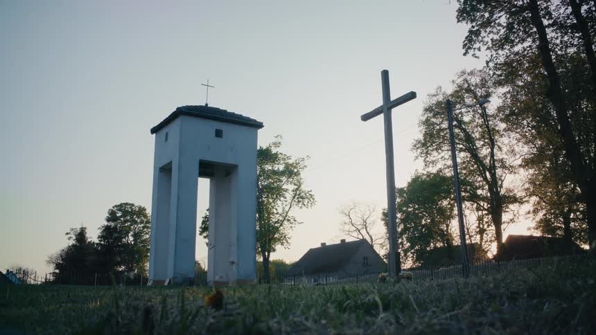 Large standing cross sits near church sitting on columns near houses under blue sky
