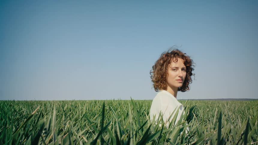A woman sits in a grassy field and then puts her arms above her head and lies down