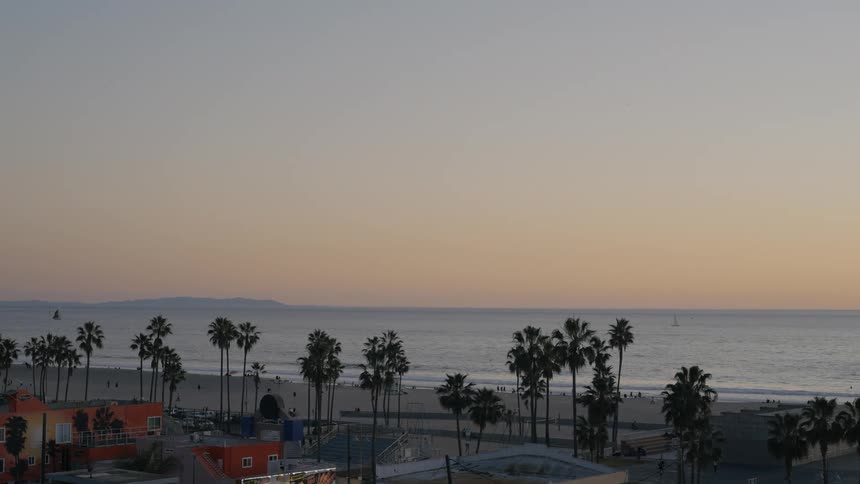 Vehicle drives along beach as people move over beach and nearby park at sunset