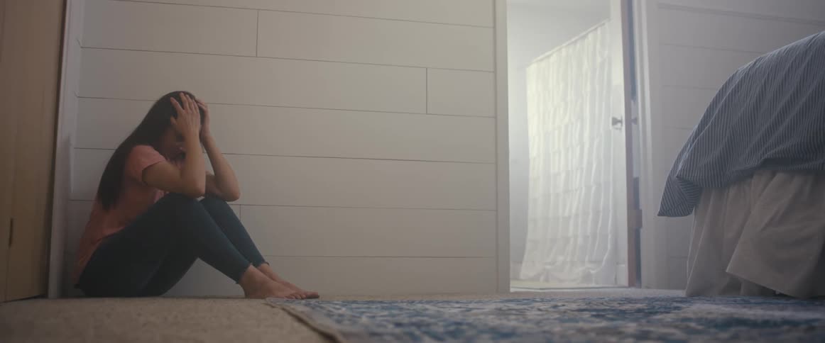 Woman weeps as she sits on the floor in the corner of a bedroom