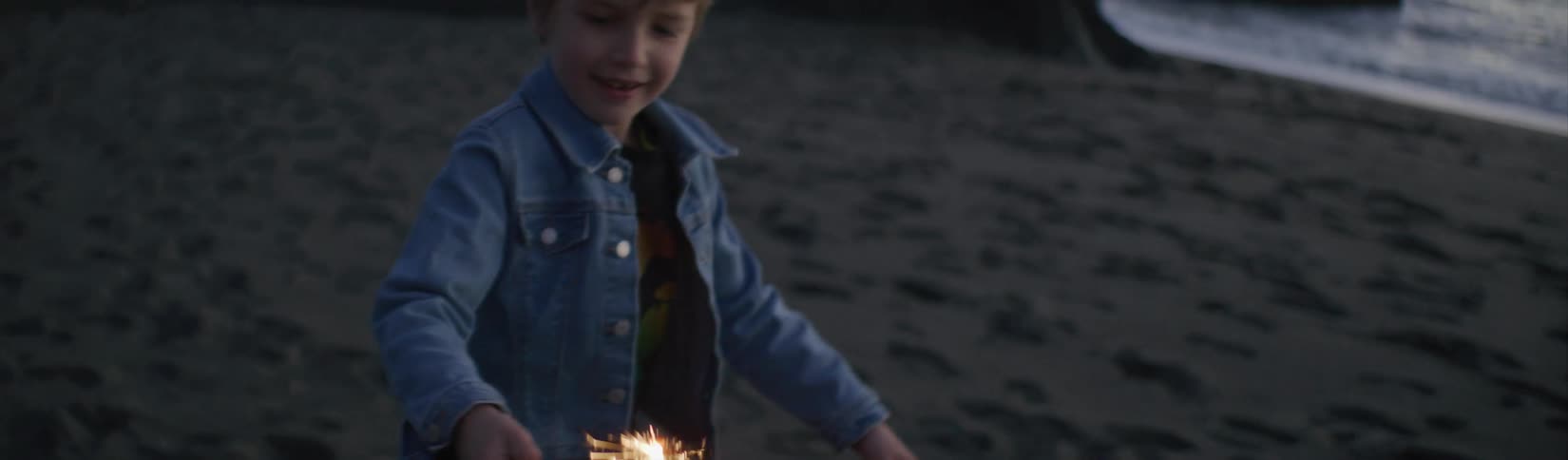 A boy laughs while running on the beach with a sparkler