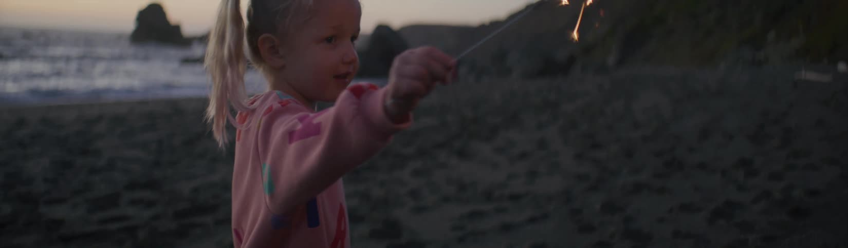 Blonde girl plays with a sparkler over the sand in the beach close to the ocean at evening