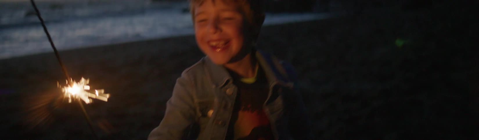 A young boy is standing on a beach holding a sparkler and smiling as he watches the sparks
