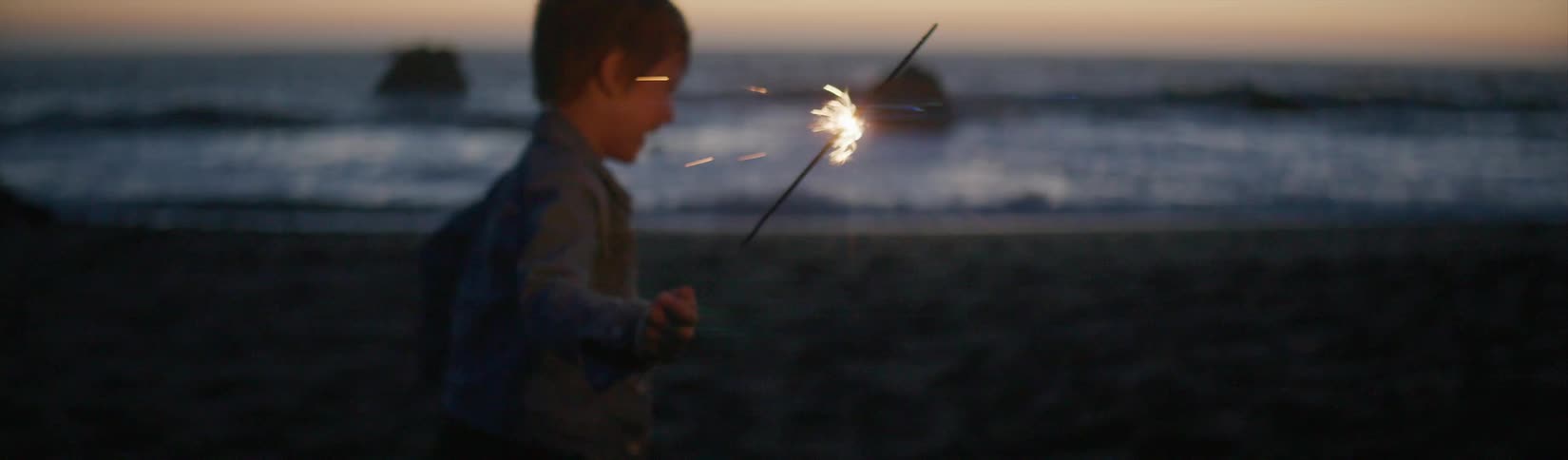 Young boy running with a sparkler near the shoreline at the beach at sunset
