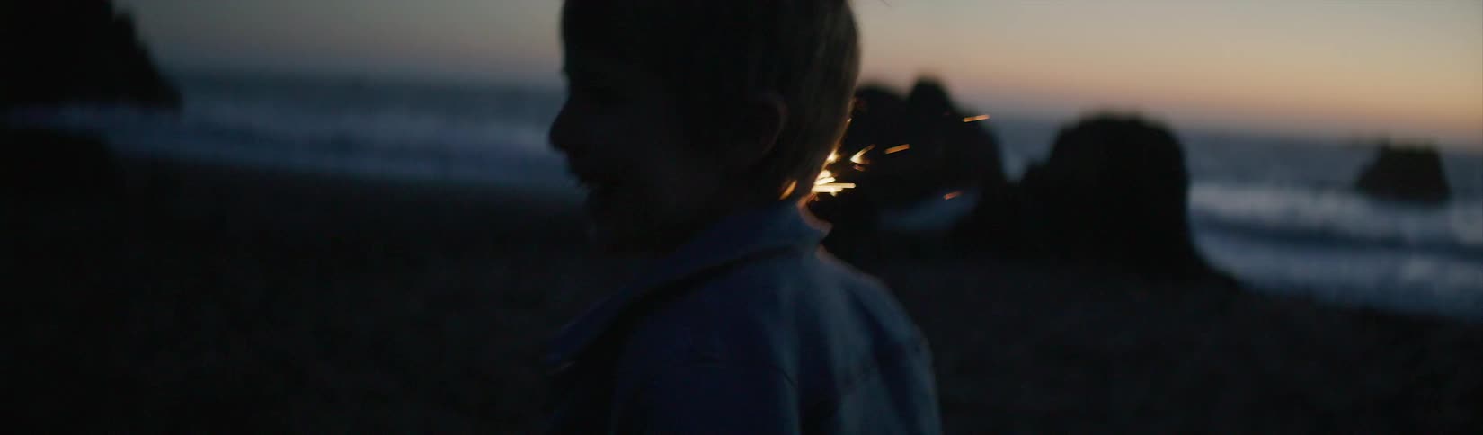 Boy runs across beach holding sparkler as afterglow remains on horizon over ocean