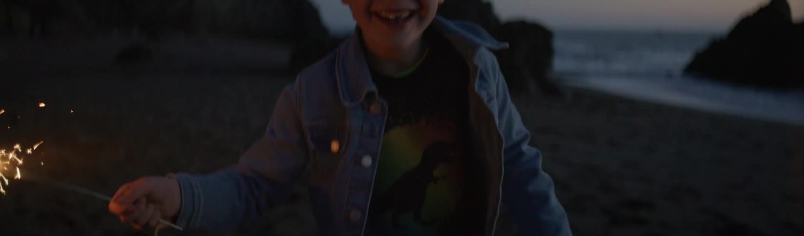 A young boy is on a beach holding a sparkler and smiles as it sparks