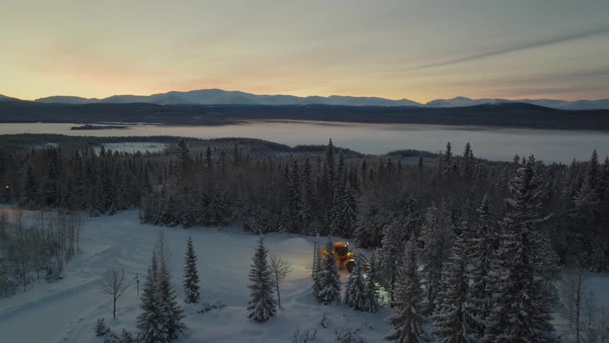 A bulldozer plows snow in a field near a forest and lake