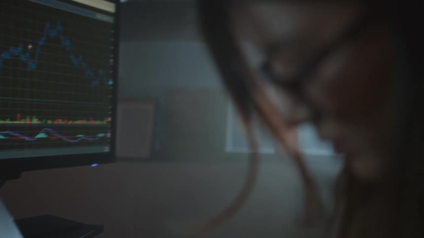 Woman sitting at desk looking at the computer and laptop screens in front of her and writing something on a notepad