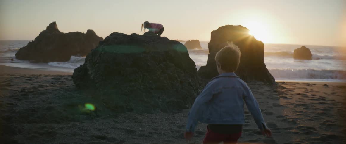 A young boy on a beach runs towards his sister who is playing on a large rock