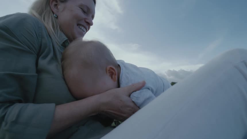 Blonde mother with green shirt and white pants holds baby sitting on grass next to father with green shirt in the field