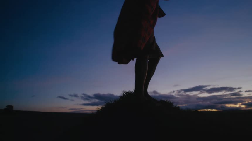 Man wearing a cloak waving in the wind climbs atop a mound at sunset