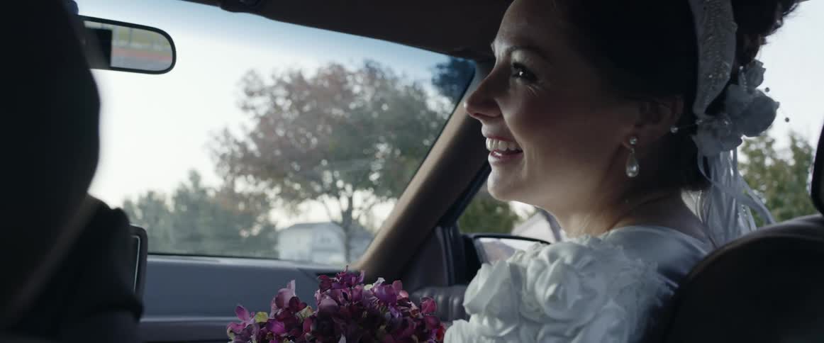 Bride in wedding dress and crown sits in carholding a bunch of flowers and lauging