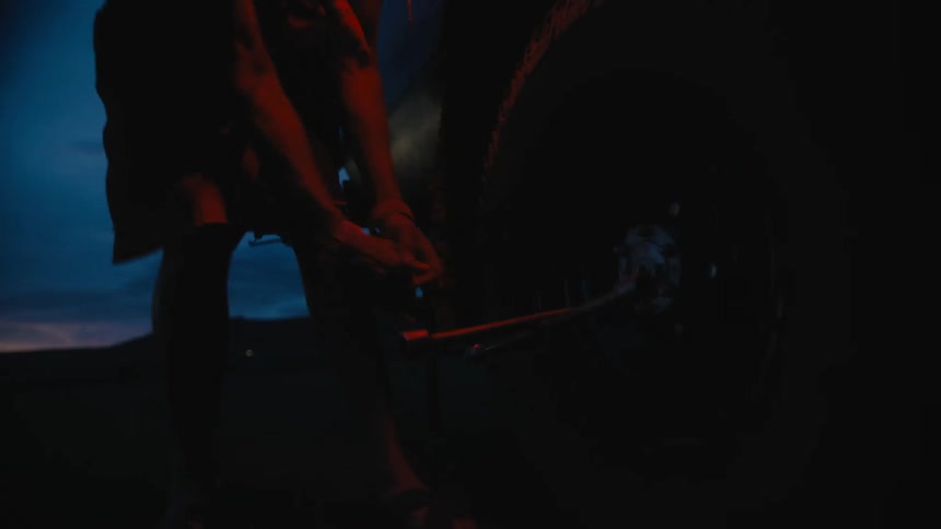 Black man using a lug wrench to change the tire of a car parked outdoors in the middle of an empty field during sunset