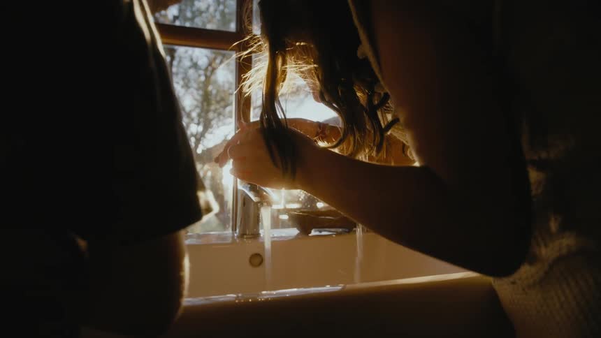 Girl with wavy hair and white shirt washes her face in the kitchen at dawn