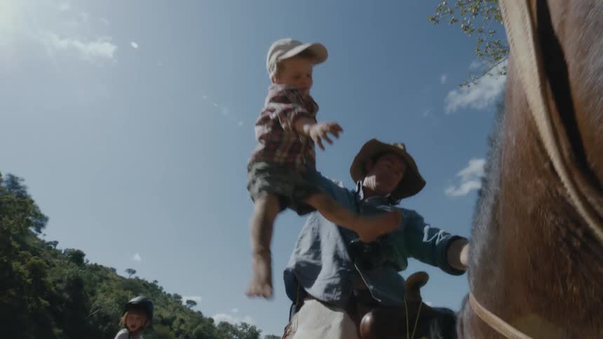 Dad pulls his toddler son up by one arm and puts him on the horse in front of him