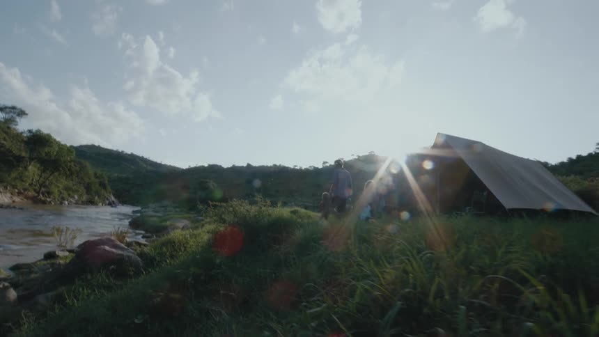 An aframe cabin is surrounded by long grass and mountains near a river