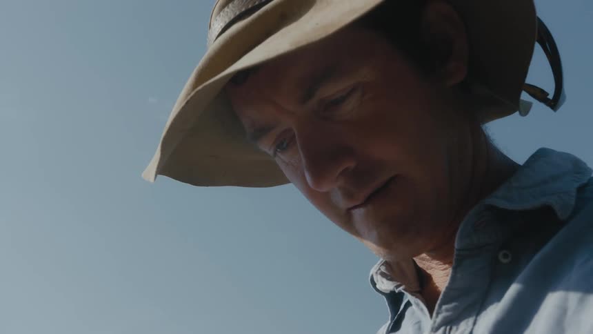 White man wearing a sun hat standing outdoors looking down and licking his lips